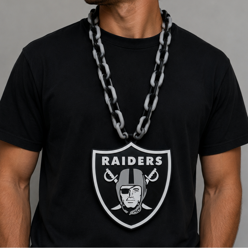 A man wearing a black Oakland Raiders t-shirt with a large Raiders shield logo on the front, a black Raiders baseball cap, and a chunky silver chain necklace, posed against a neutral gray background.