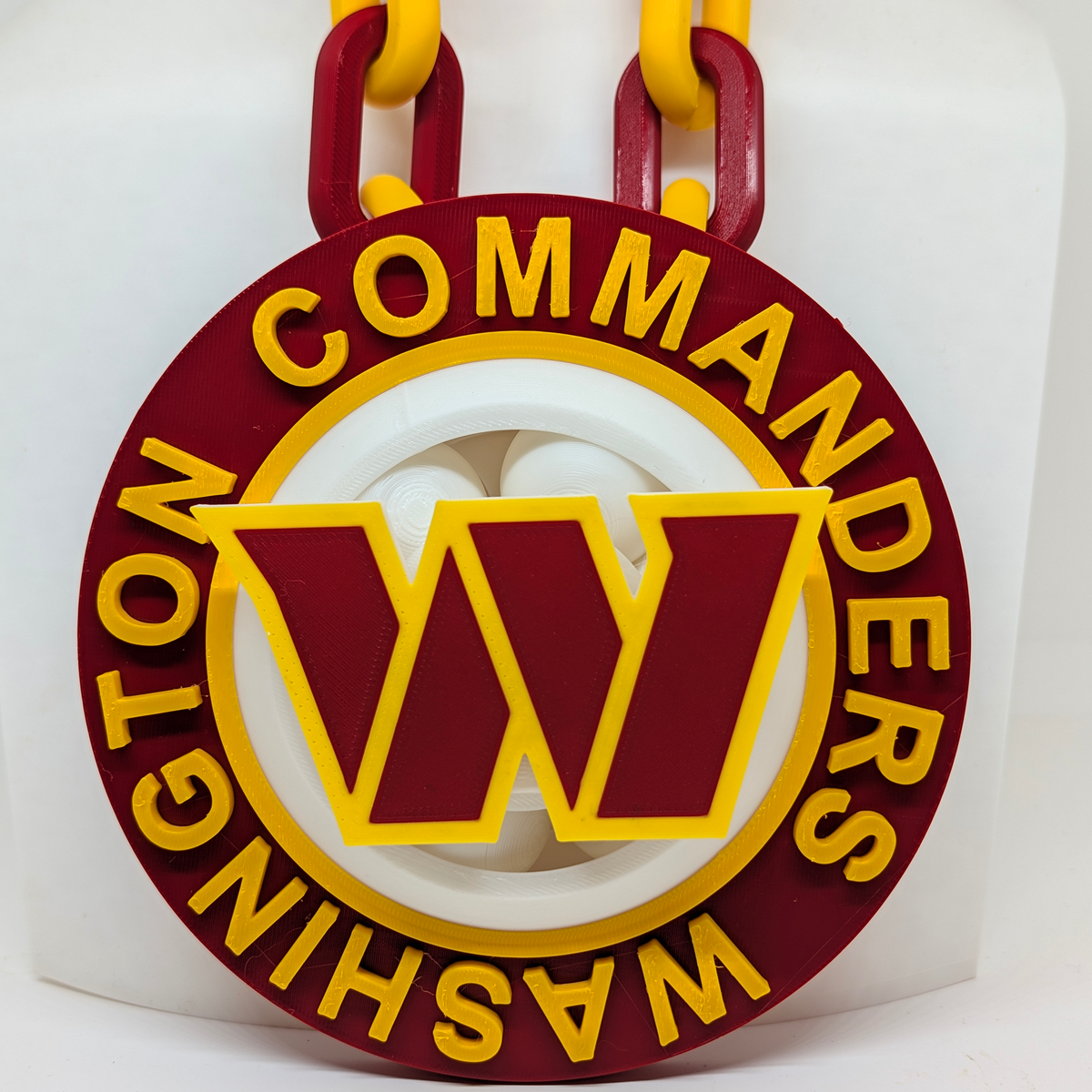 A Washington Commanders themed circular pendant or medallion with a large yellow 'W' logo in the center, surrounded by yellow text reading 'WASHINGTON COMMANDERS' on a dark red background, hanging from thick yellow and dark red chain links against a white backdrop.