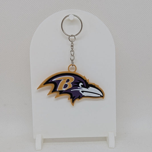 Ravens Keychain with a logo on a white stand against a light gray background